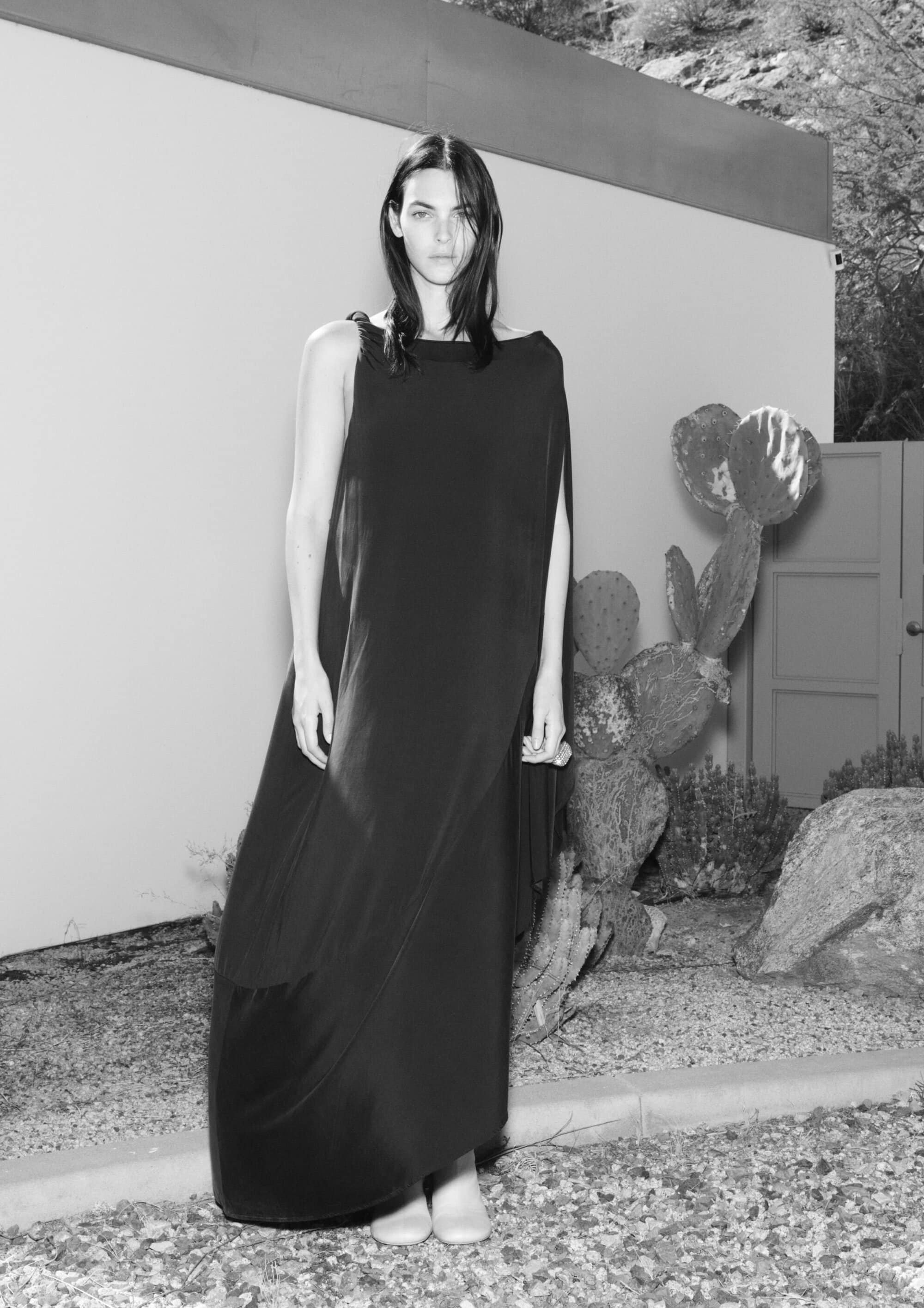 Black and white fashion shot of a woman in a long gown, posing outdoors near a house and cacti, with soft lighting and desert landscape.