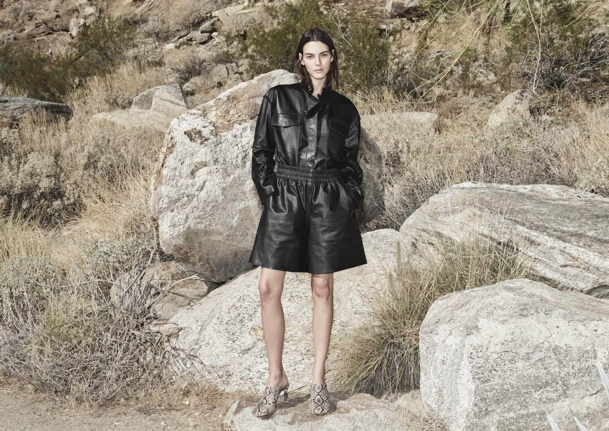 Woman in black leather jacket and shorts stands in desert landscape with rocks, wearing snakeskin shoes, creating a fashion editorial.