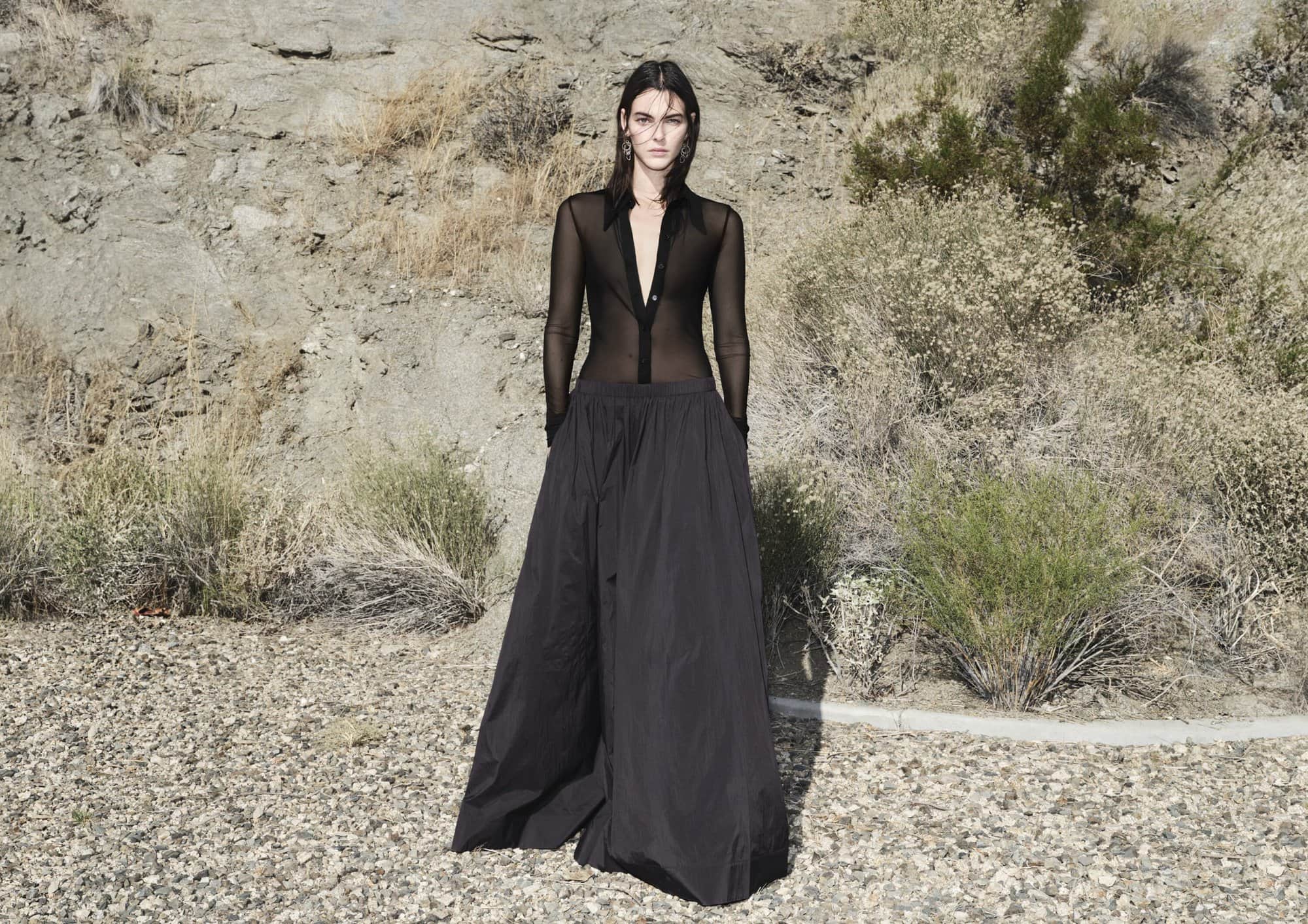 An adult woman poses in a sheer black top and long black skirt in a desert environment with a rocky hillside and sparse vegetation.