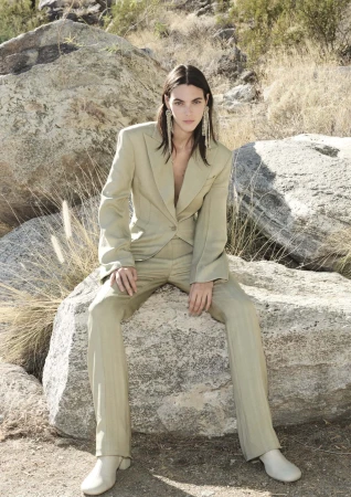 A slender person with long dark hair, wearing a light-beige suit, sits on a rock in a desert landscape with sparse vegetation and arid hills.