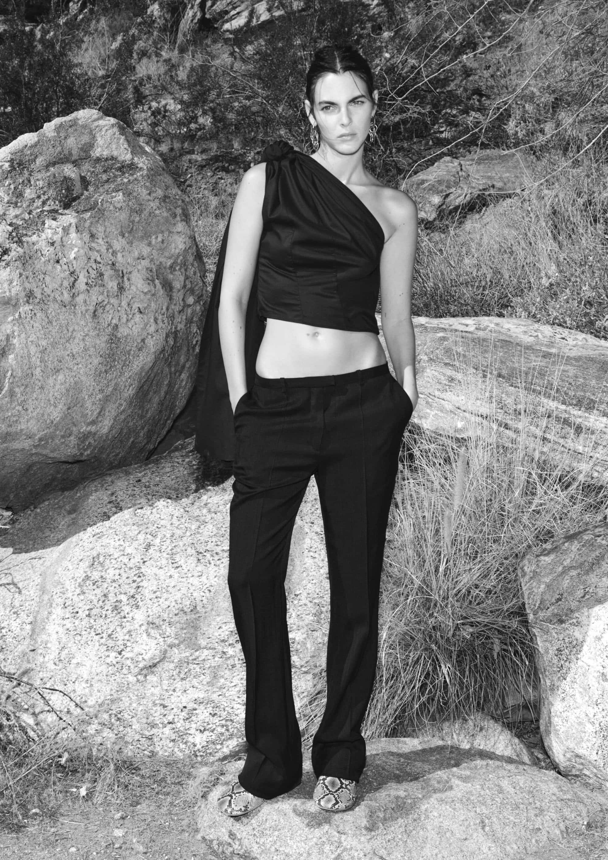 Monochrome fashion shot of a woman in a one-shoulder top and trousers, posing among rocks and sparse vegetation in a natural outdoor setting.