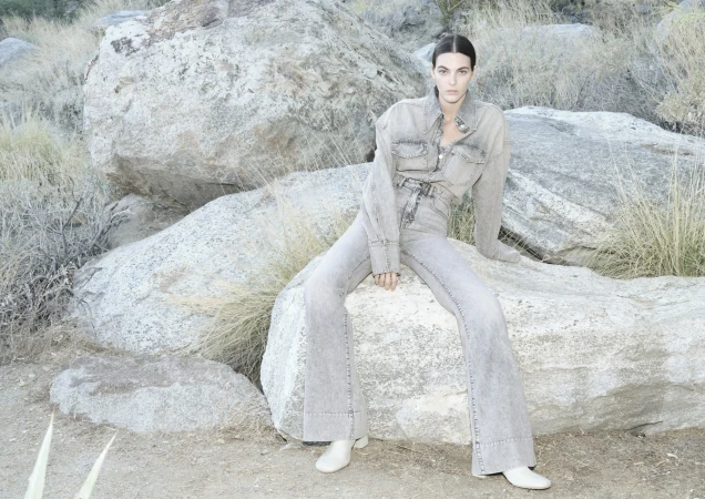 A fashionable woman in denim sits among desert rocks, wearing a stylish light denim shirt and wide-leg jeans, paired with white shoes.