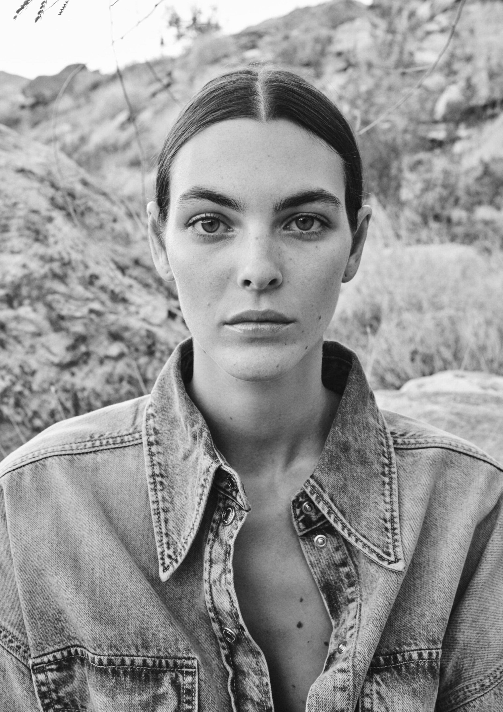 Monochrome portrait of a woman with slicked-back hair wearing a denim jacket, posing in an outdoor, rocky environment.