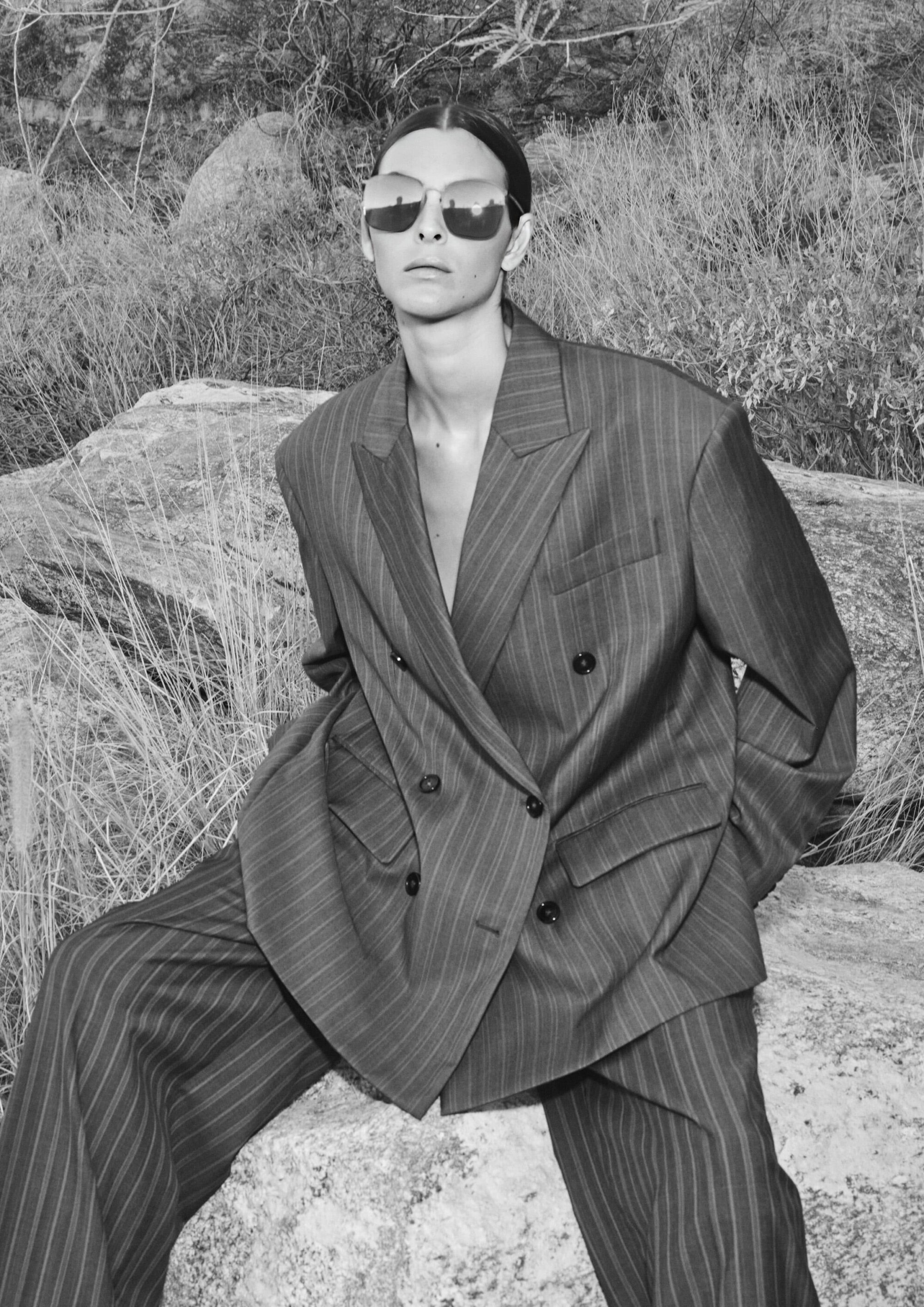 Black and white outdoor fashion shot of a stylish woman in a striped suit and mirrored sunglasses sitting on a rock with dry grass.