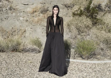 Vittoria Ceretti wearing a black sheer blouse and long dark skirt standing outdoors before a rocky arid hillside for H&M.