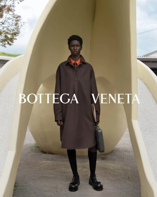 Fashion shot featuring a model in a brown dress, black socks and loafers, holding a bag, posing in front of a white structure.