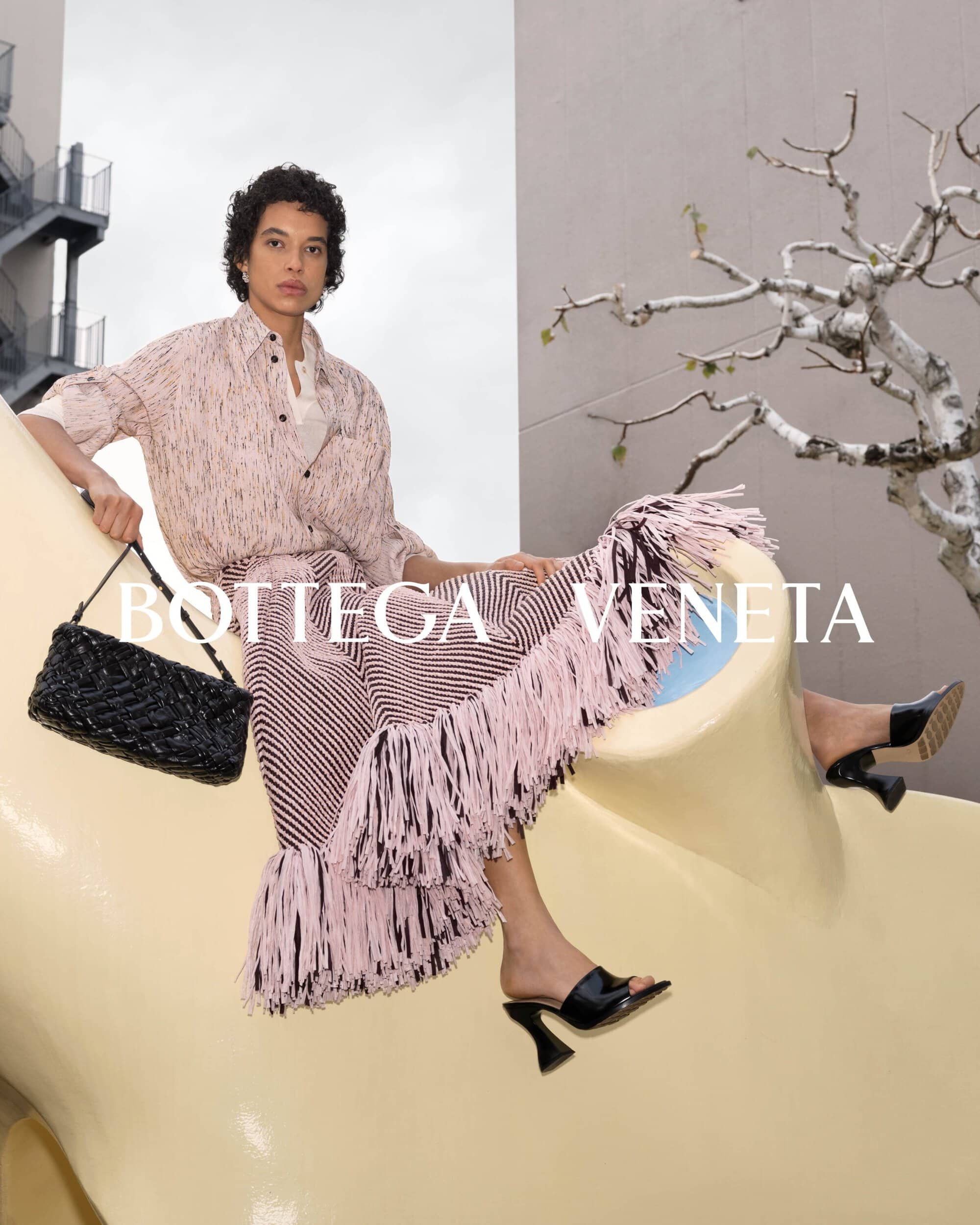 A woman wearing a patterned skirt, blouse, and black high heels is sitting and posing with a black woven bag in an outdoor environment.
