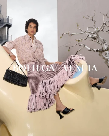 A woman wearing a patterned skirt, blouse, and black high heels is sitting and posing with a black woven bag in an outdoor environment.