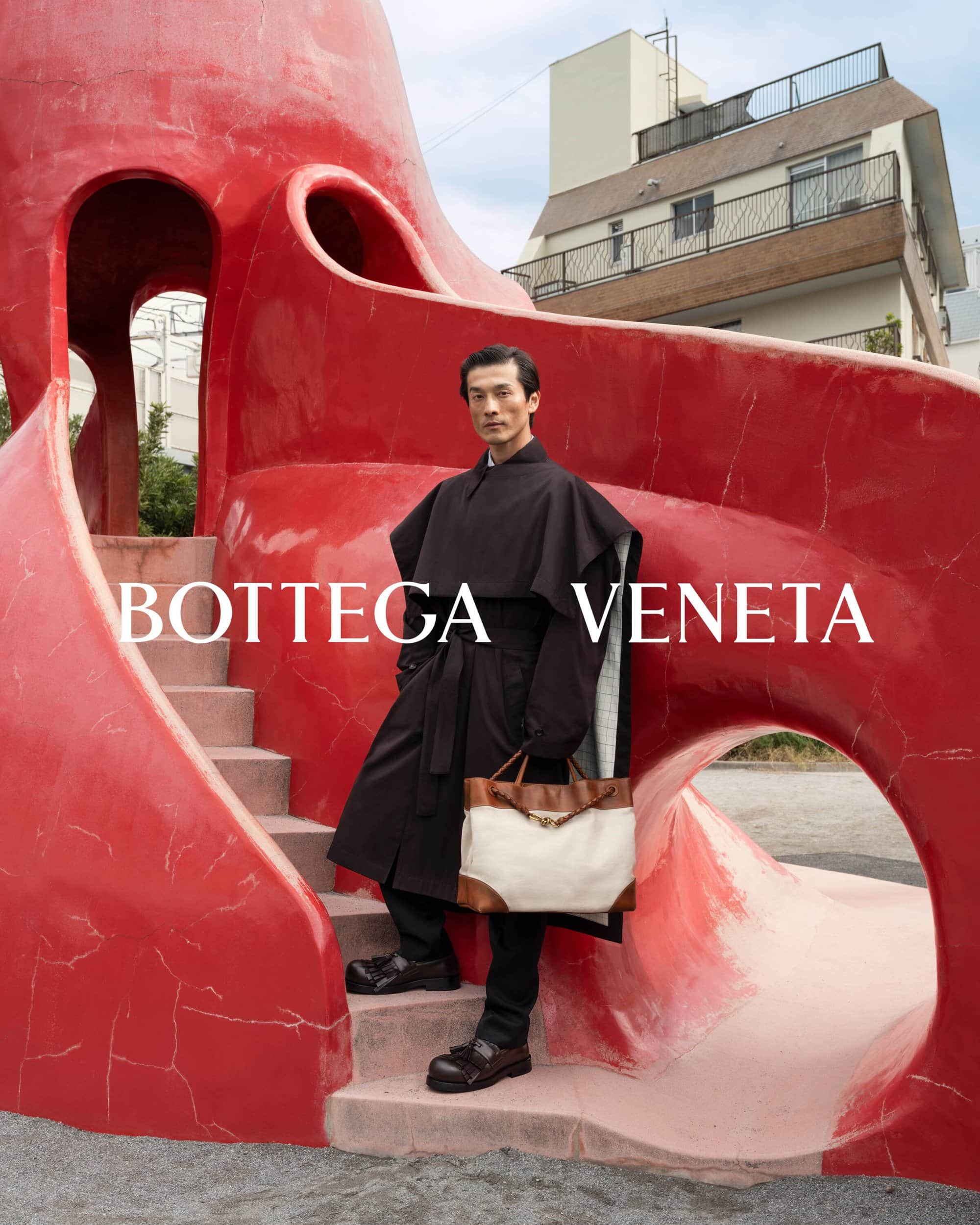 Adult male in outdoor fashion shot stands near red sculpture wearing a long coat and carrying a tan and white bag. 