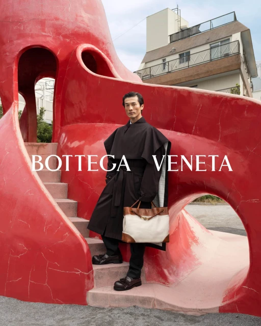 Adult male in outdoor fashion shot stands near red sculpture wearing a long coat and carrying a tan and white bag. 