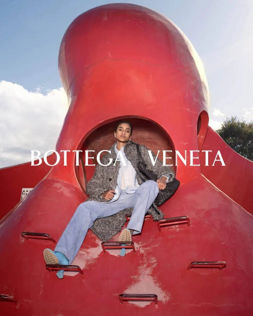 Fashion shot of woman in coat and jeans sitting on a large red octopus sculpture against a blue sky for Bottega Veneta campaign.