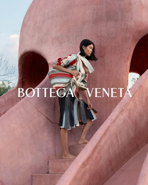 Fashion shot of a woman climbing pink steps wearing a striped top, dark skirt, and carrying a handbag, with a building in the background.