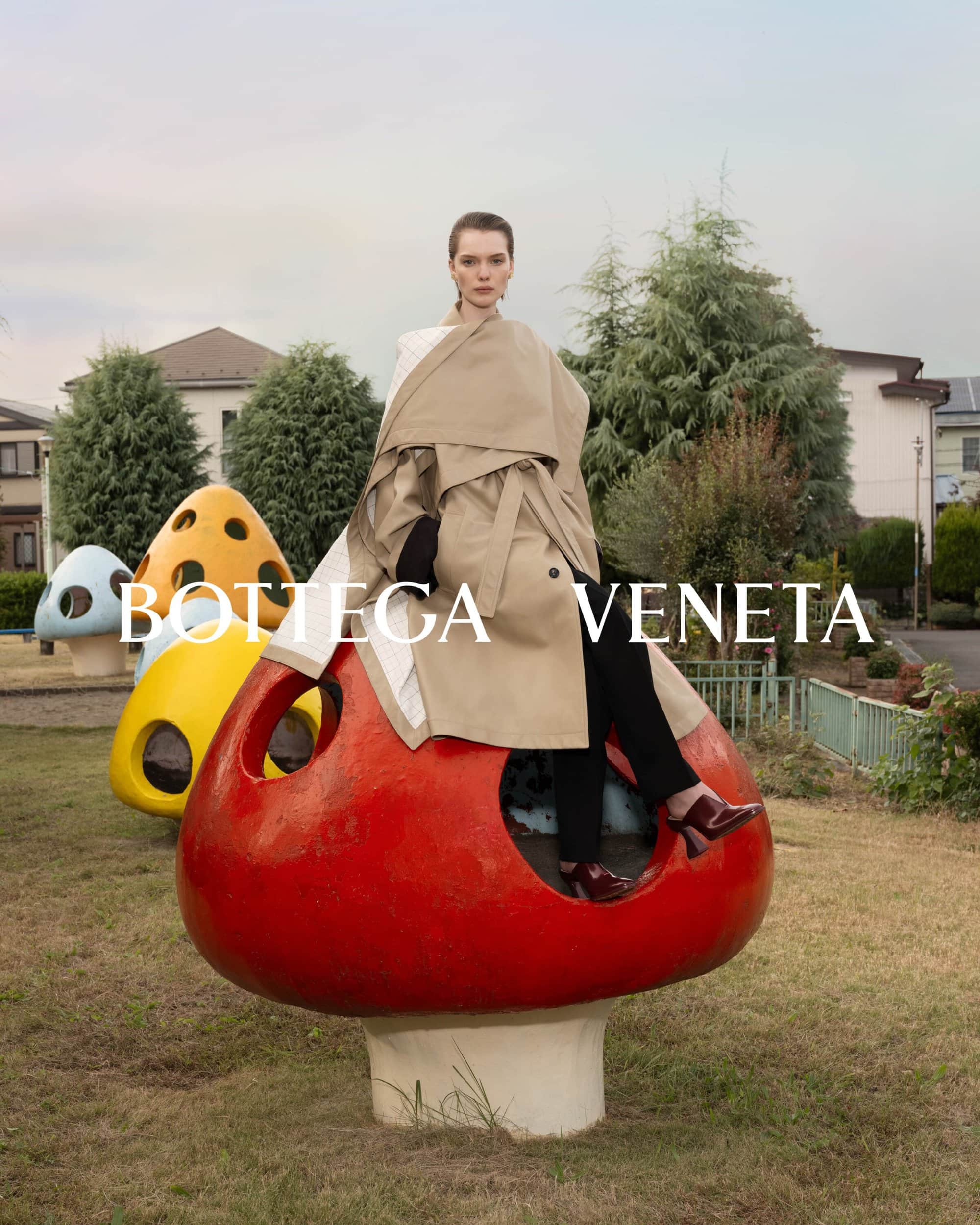 Fashion shot of a woman in a tan trench coat sitting on a red mushroom prop in a grassy park with buildings and trees in the background.