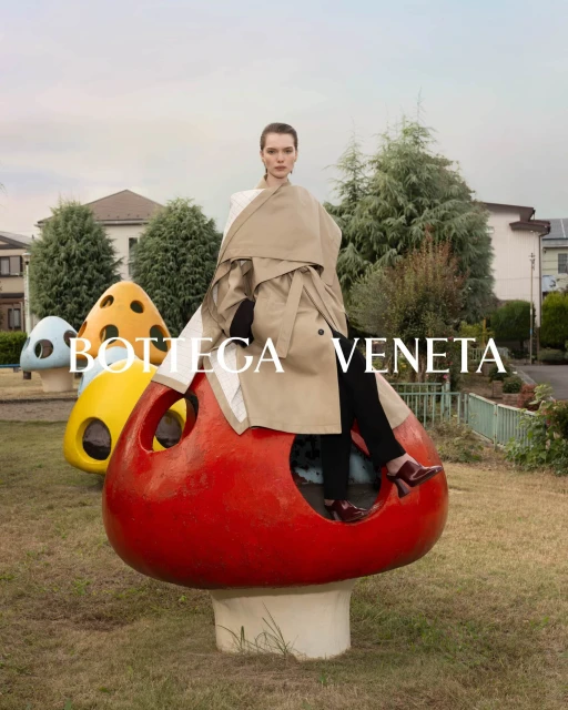 Fashion shot of a woman in a tan trench coat sitting on a red mushroom prop in a grassy park with buildings and trees in the background.