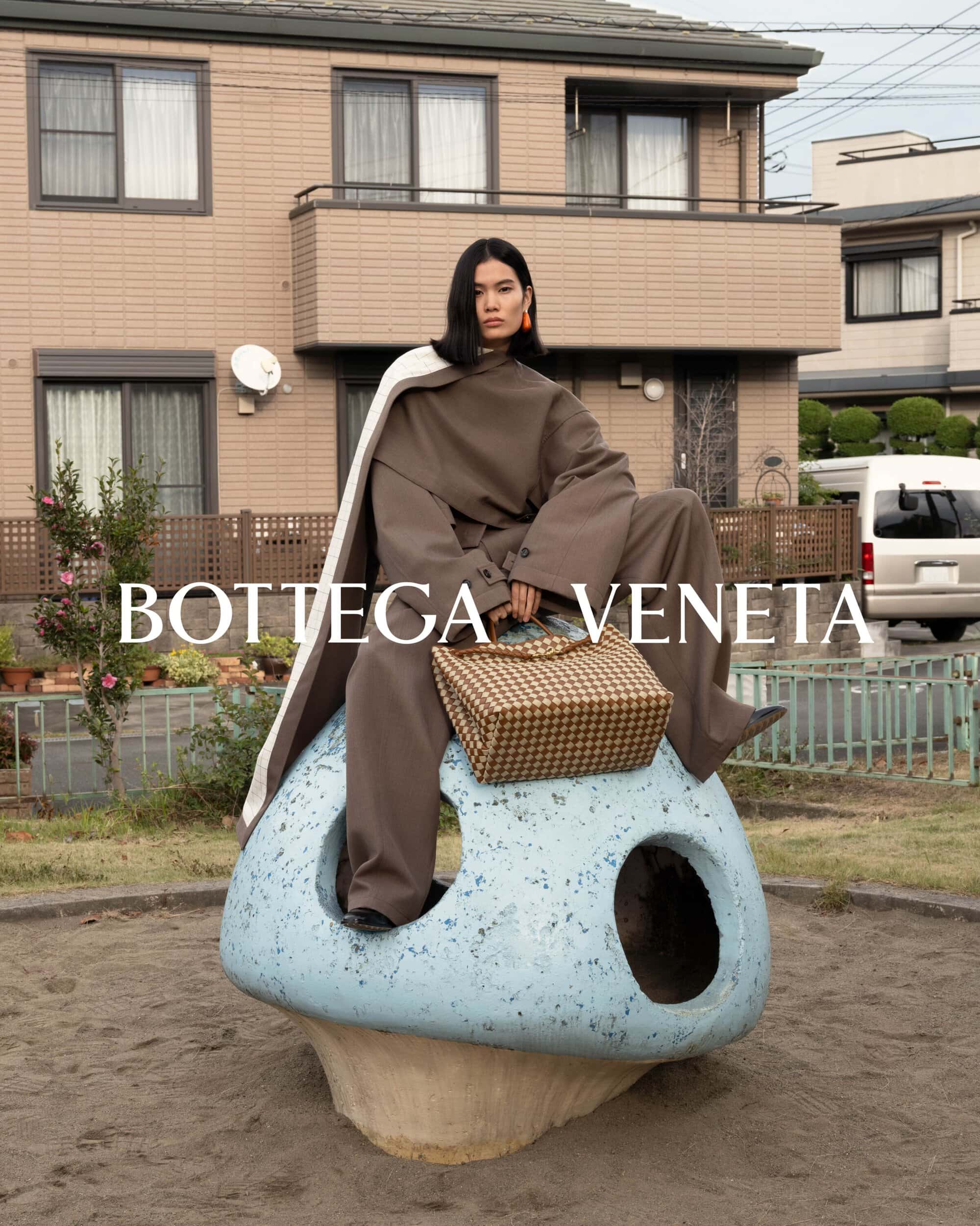 Fashion shot of a woman in olive-green pantsuit and a white cape, seated on a blue mushroom sculpture with a woven handbag.