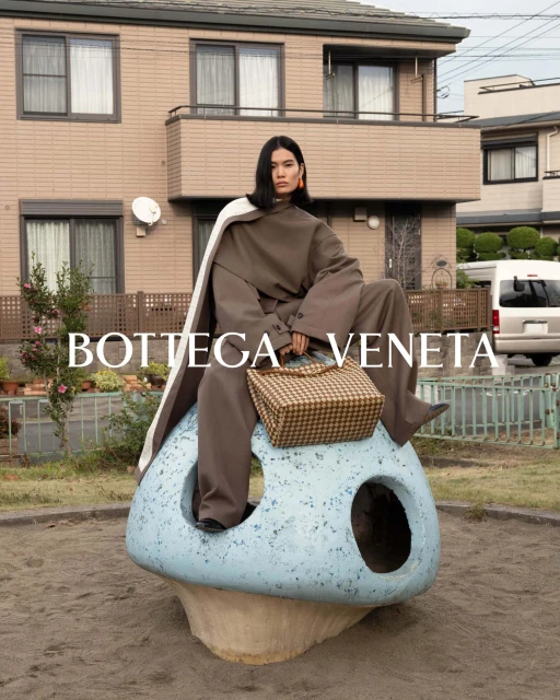 Fashion shot of a woman in olive-green pantsuit and a white cape, seated on a blue mushroom sculpture with a woven handbag.
