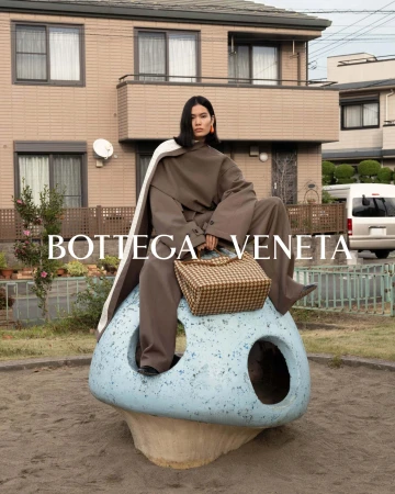 Fashion shot of a woman in olive-green pantsuit and a white cape, seated on a blue mushroom sculpture with a woven handbag.