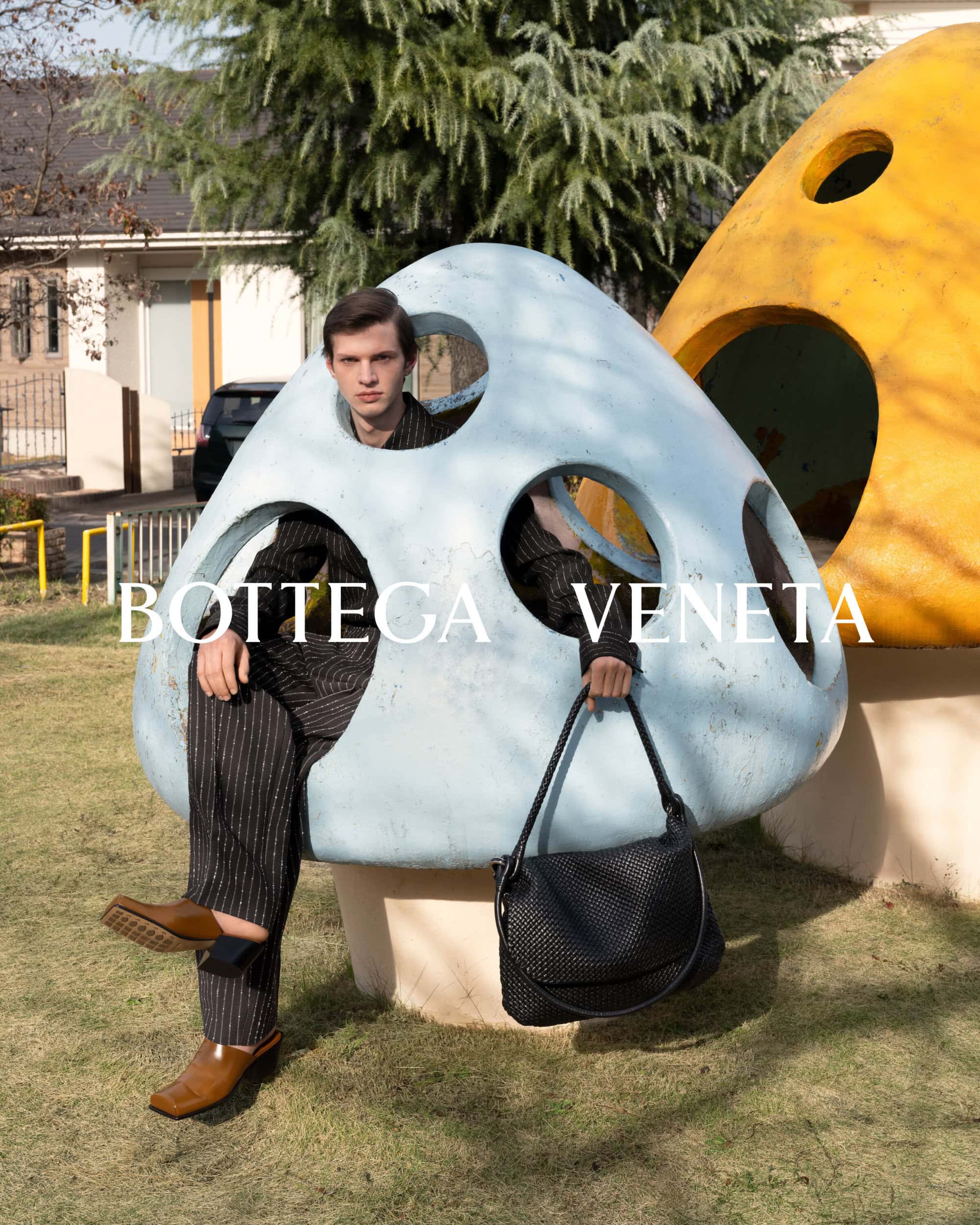 An adult male fashion model wearing a striped suit and loafers is seated on a mushroom statue in a park, holding a black Bottega Veneta bag.