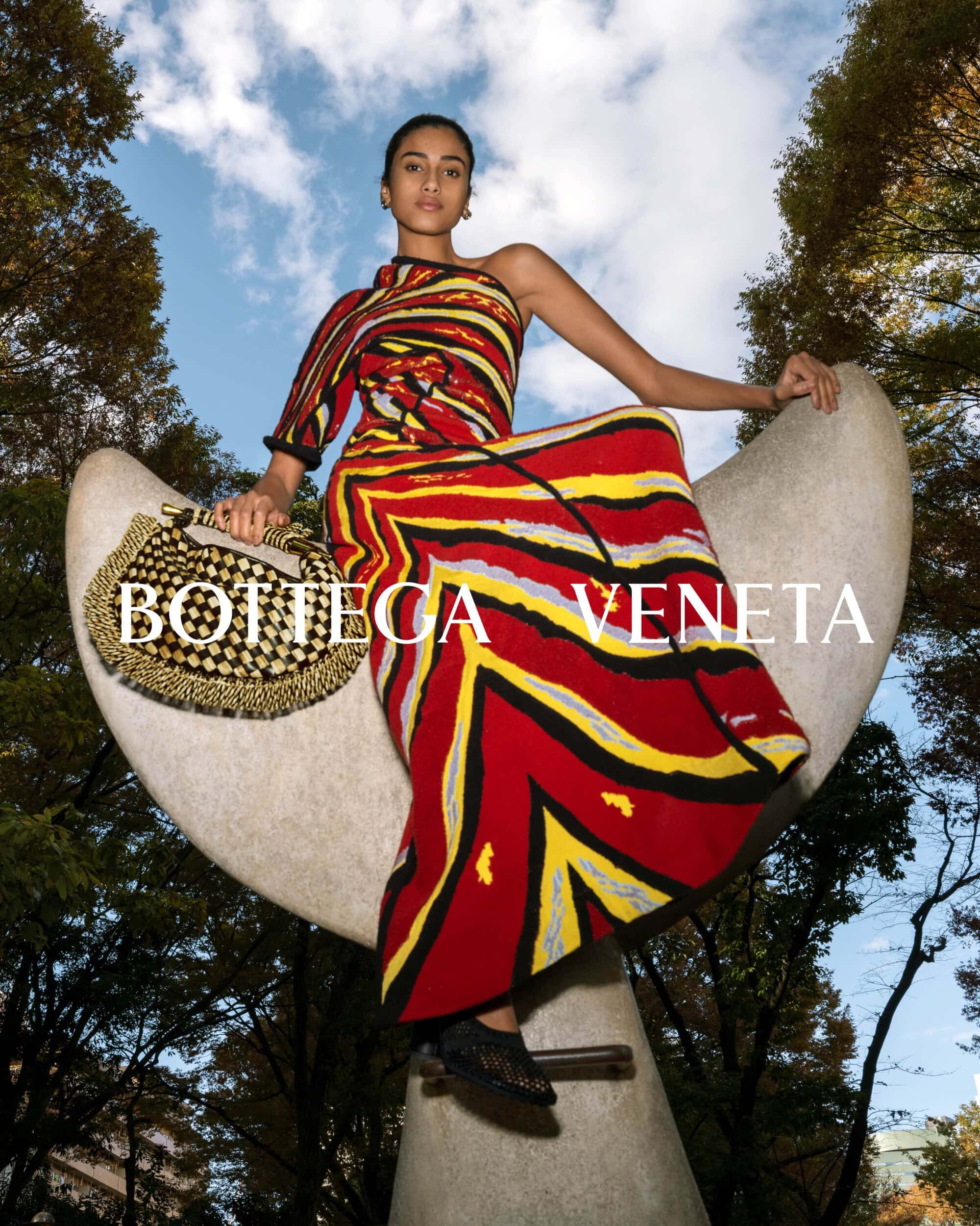 Adult female in a red and yellow dress with a black bag poses on a gray stone structure with trees and sky in the background.
