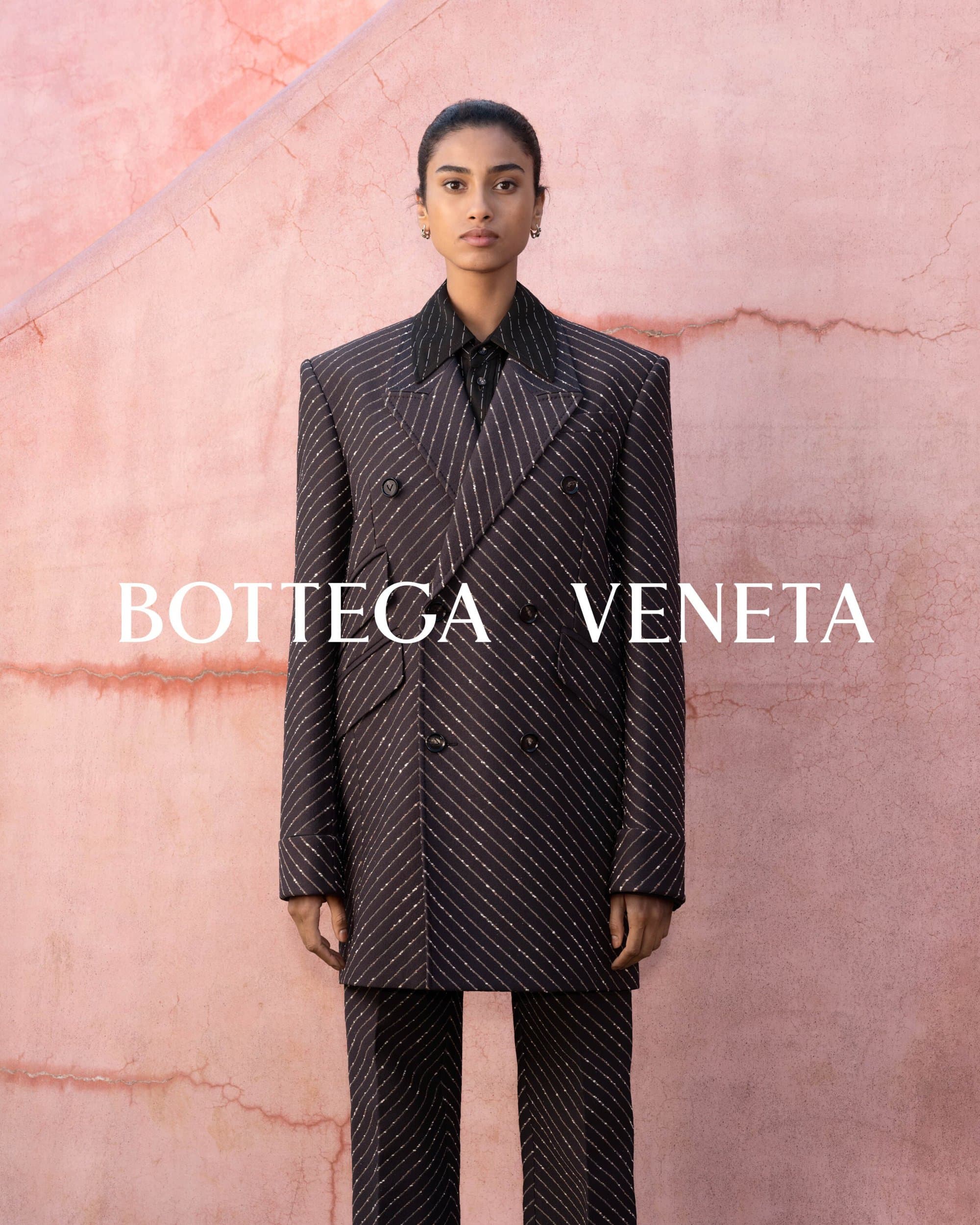 A South Asian woman in a pinstripe suit stands against a pink textured wall. Bottega Veneta logo is layered over the image in white.
