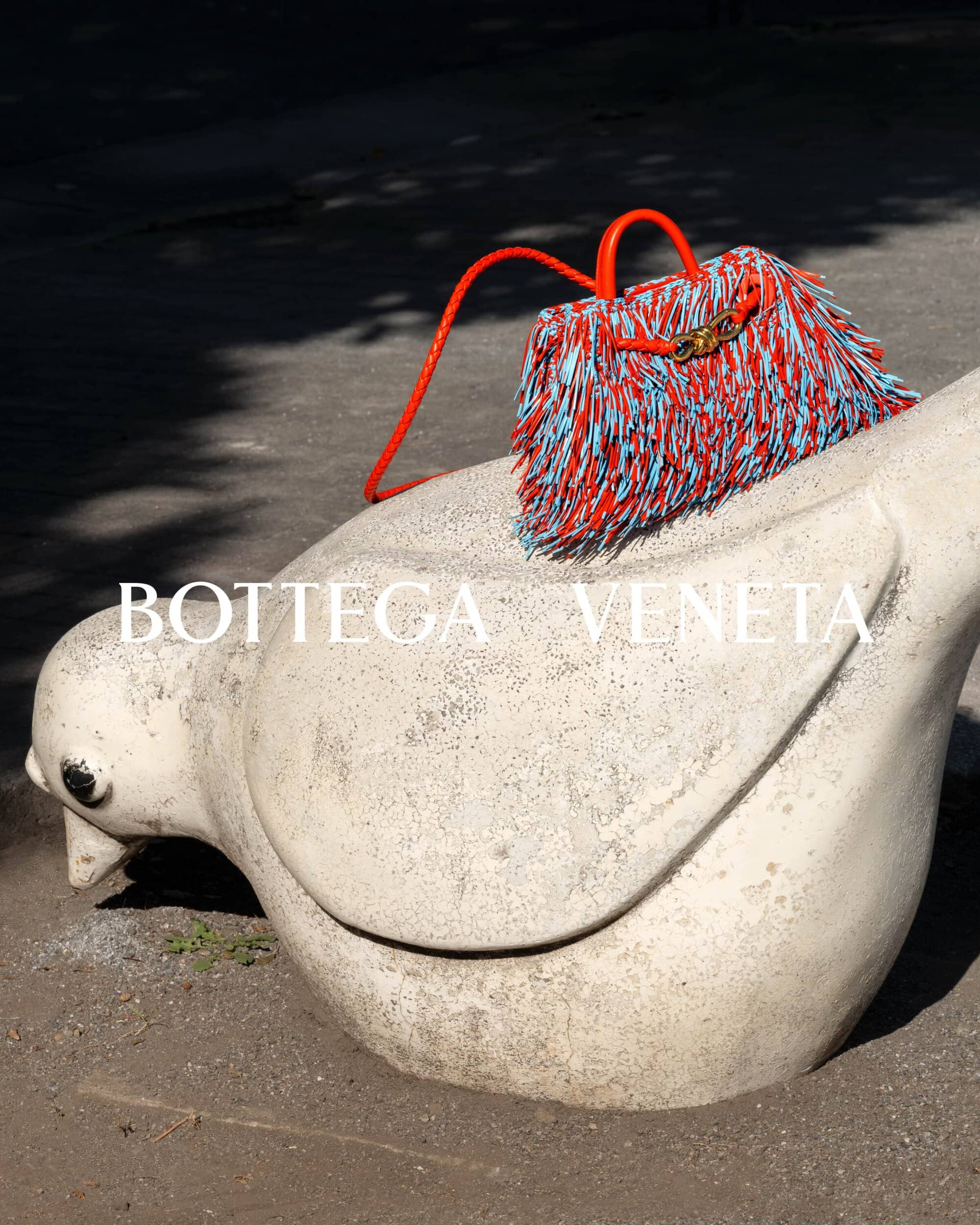 Orange and blue fringed Bottega Veneta handbag sitting on a weathered white stone bird sculpture outdoors in bright sunlight.