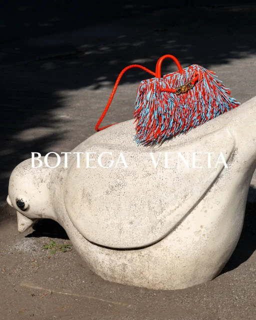 Orange and blue fringed Bottega Veneta handbag sitting on a weathered white stone bird sculpture outdoors in bright sunlight.