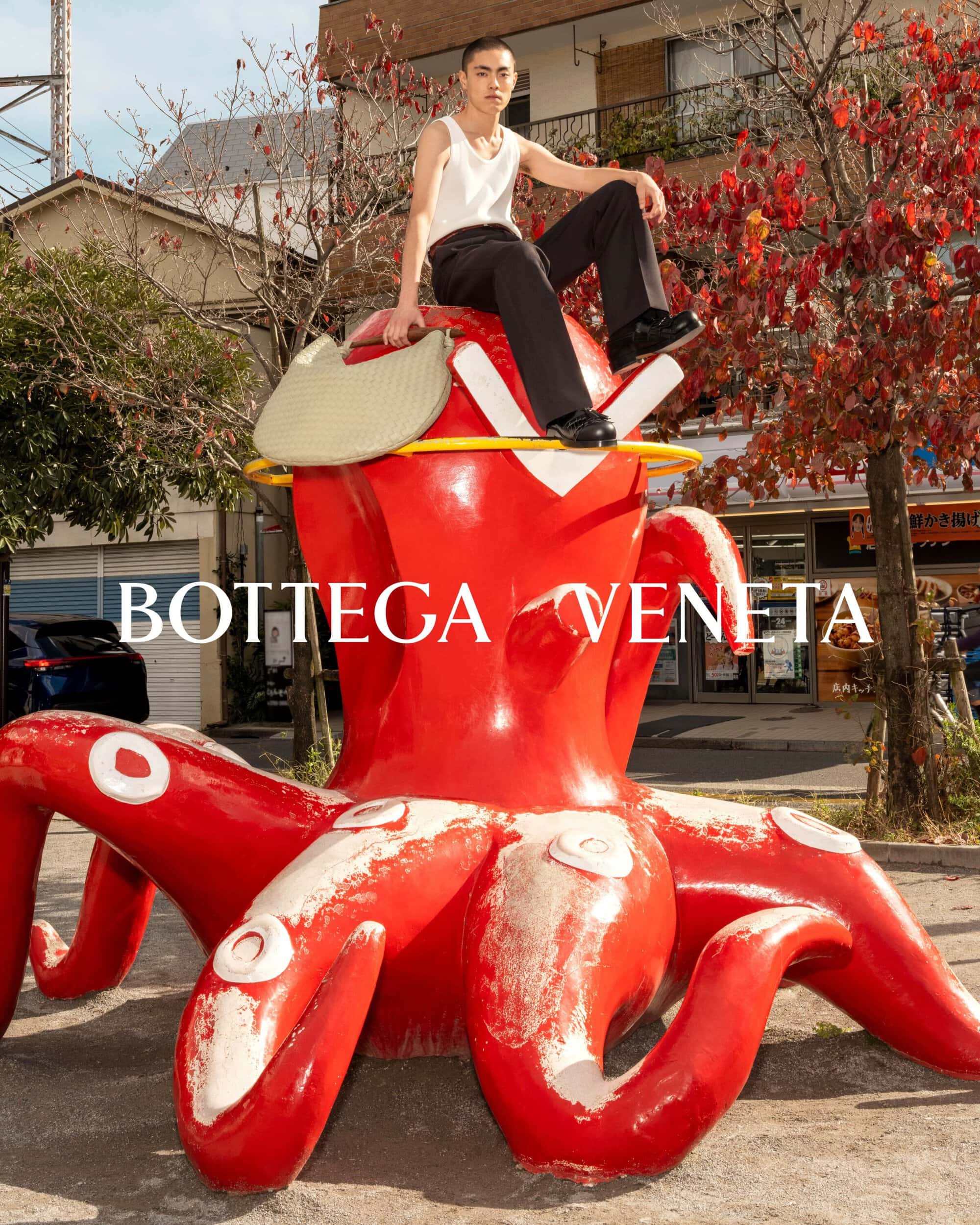 A person sits on a large red octopus sculpture holding a woven tote bag, pictured outdoors with Bottega Veneta logo, buildings, and trees.