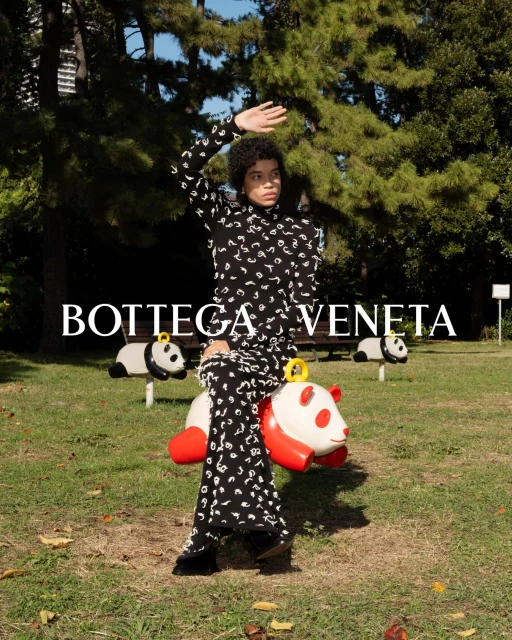 Woman in patterned outfit poses on playground ride in park setting with a large designer logo in the foreground.