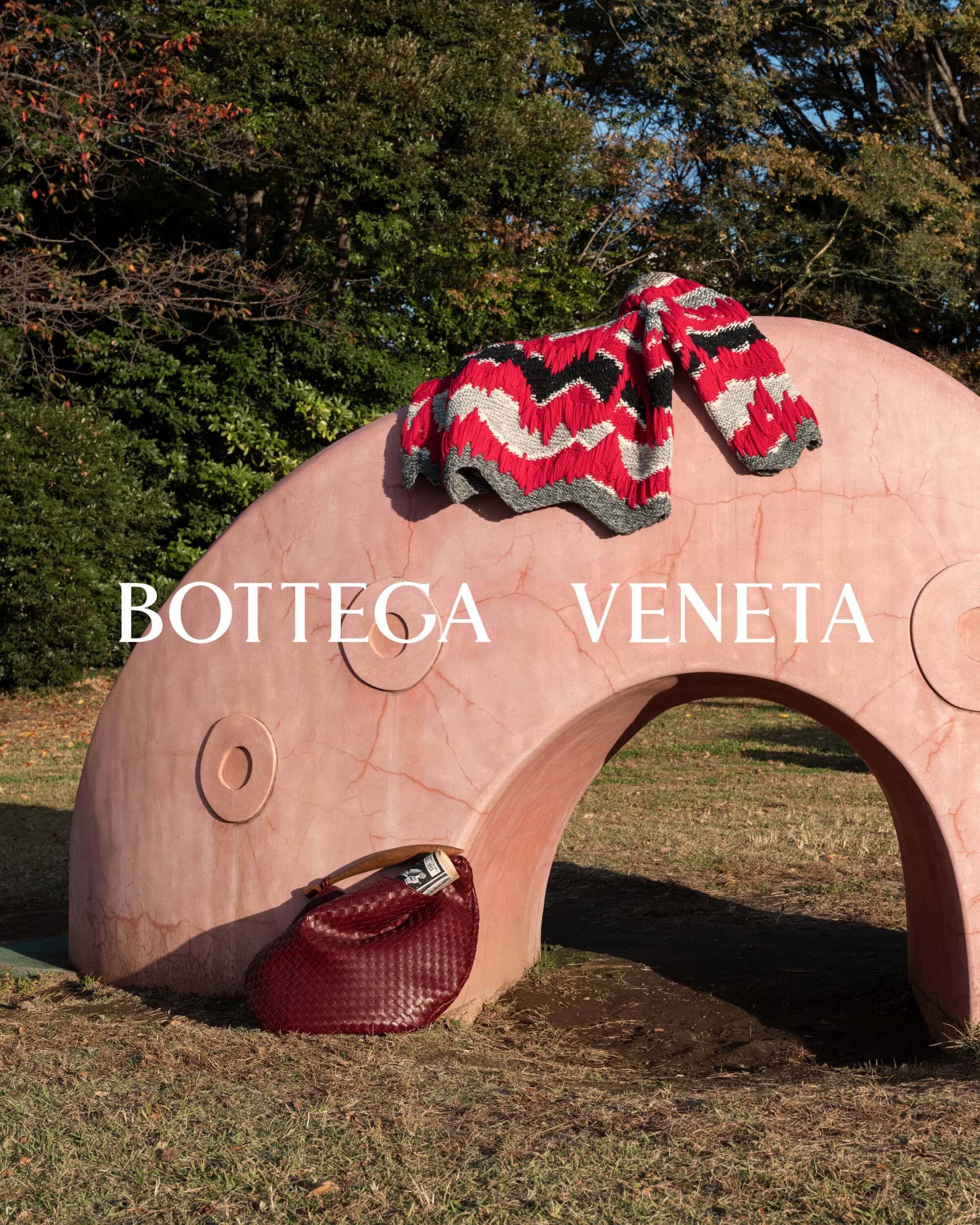 Bottega Veneta: Red patterned sweater sits atop a pink archway with a red woven handbag on grass outdoors with trees.