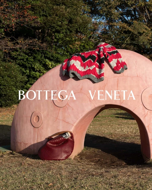 Bottega Veneta: Red patterned sweater sits atop a pink archway with a red woven handbag on grass outdoors with trees.