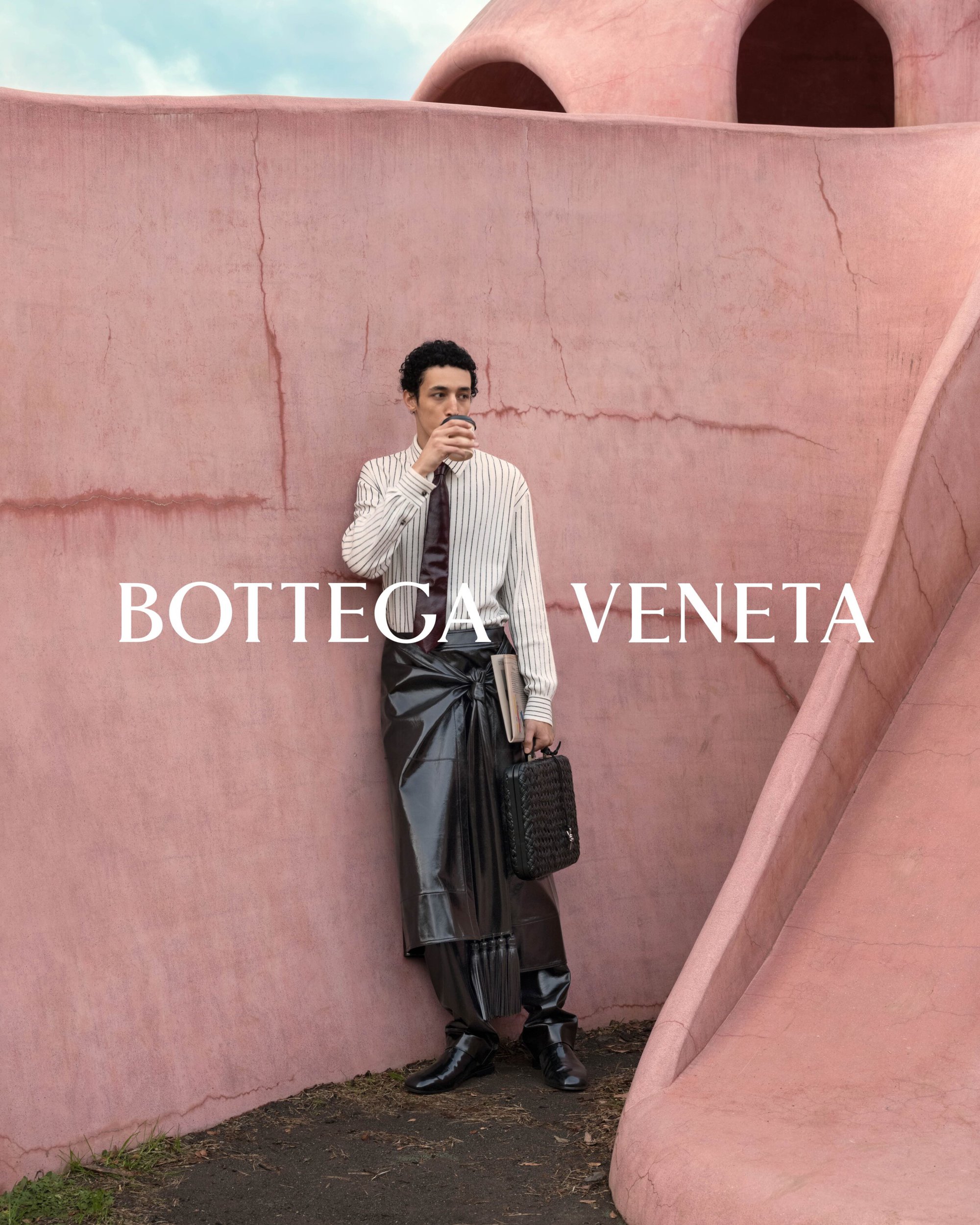 A man in a striped shirt and black skirt drinks from a cup outdoors with a black bag, posing against a pink wall and slide.