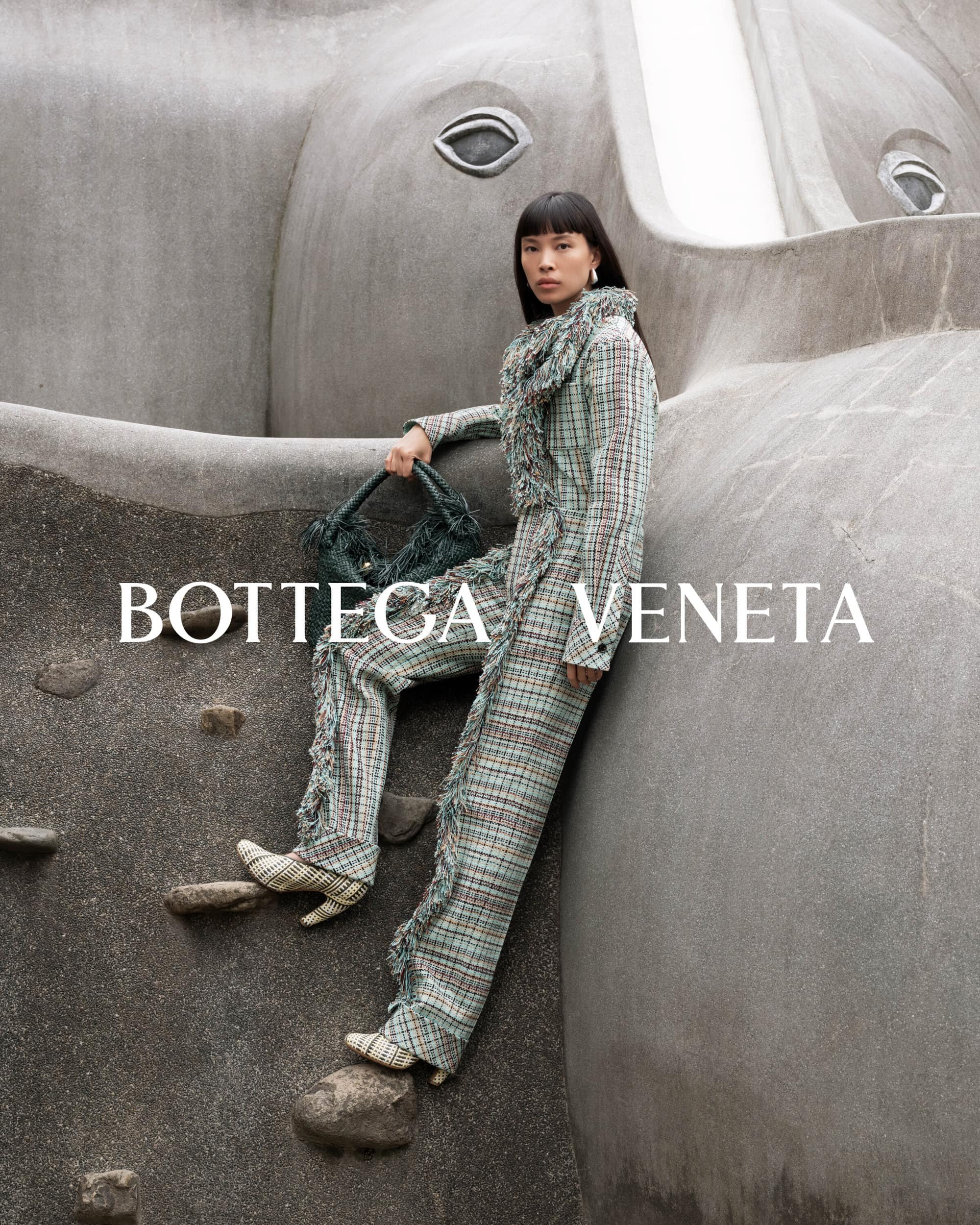 Fashionable Asian woman poses with handbag next to a gray sculpture in a Bottega Veneta advert featuring unique ready-to-wear.