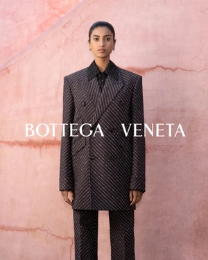 Model wearing a dark pinstripe double-breasted suit standing against a textured pink wall with Bottega Veneta text.