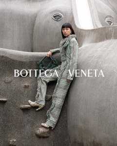 Model in plaid fringe trousers and matching top holding a dark woven handbag against a sculptural grey stone structure.