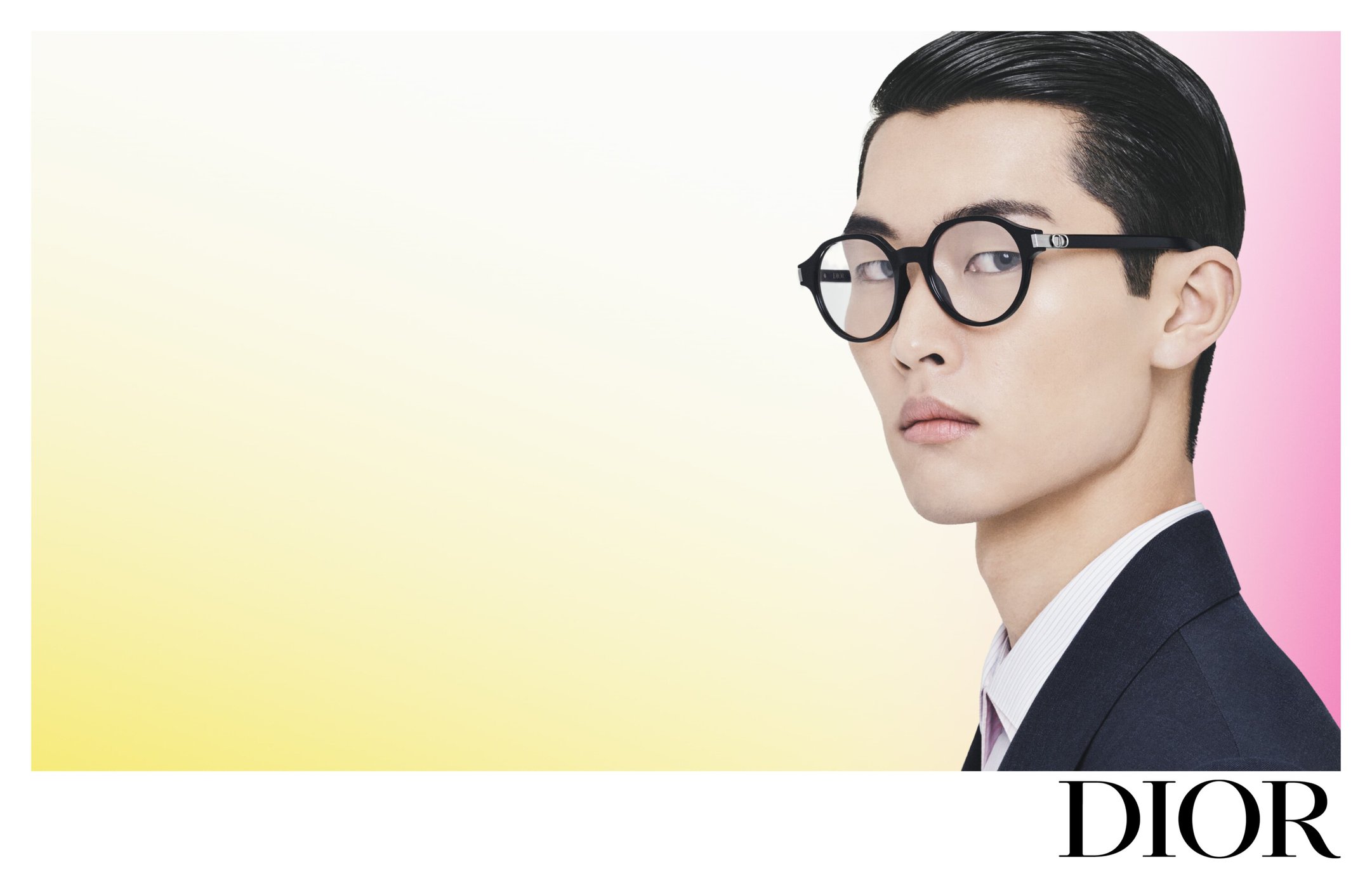 Close-up studio shot of an Asian male model wearing round black-rimmed eyeglasses, a dark blazer, and a white collared shirt. Dior logo in corner.