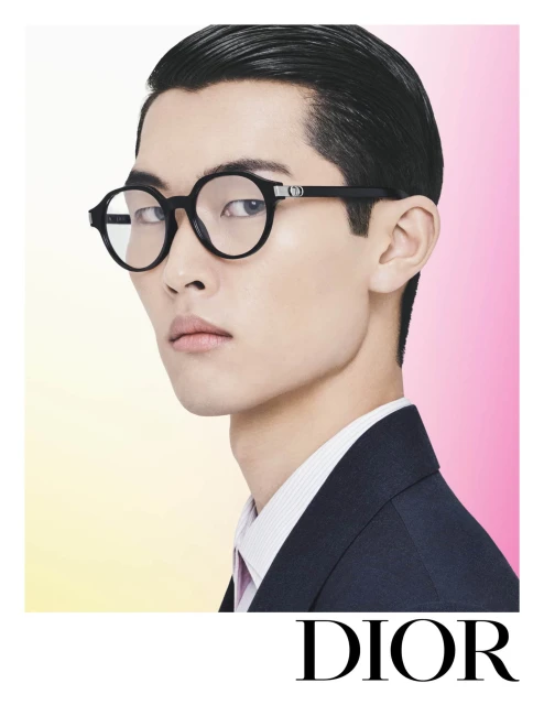Close-up of an Asian male model wearing black framed glasses and a dark blue suit jacket against a graduated pink background.