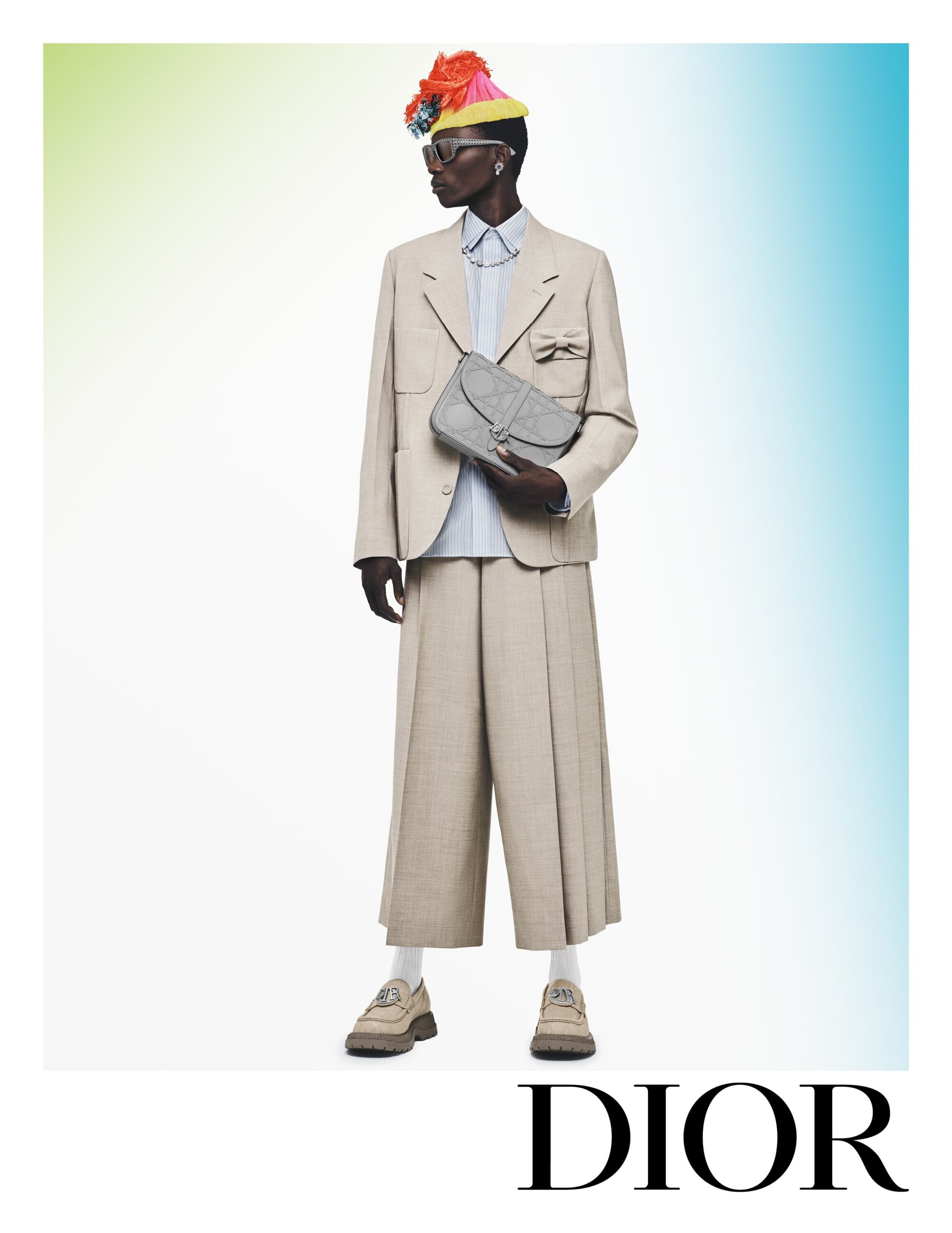 Fashion editorial shot of an African-descent adult posing in a suit with a handbag, hat, and platform shoes on a light gradient backdrop.