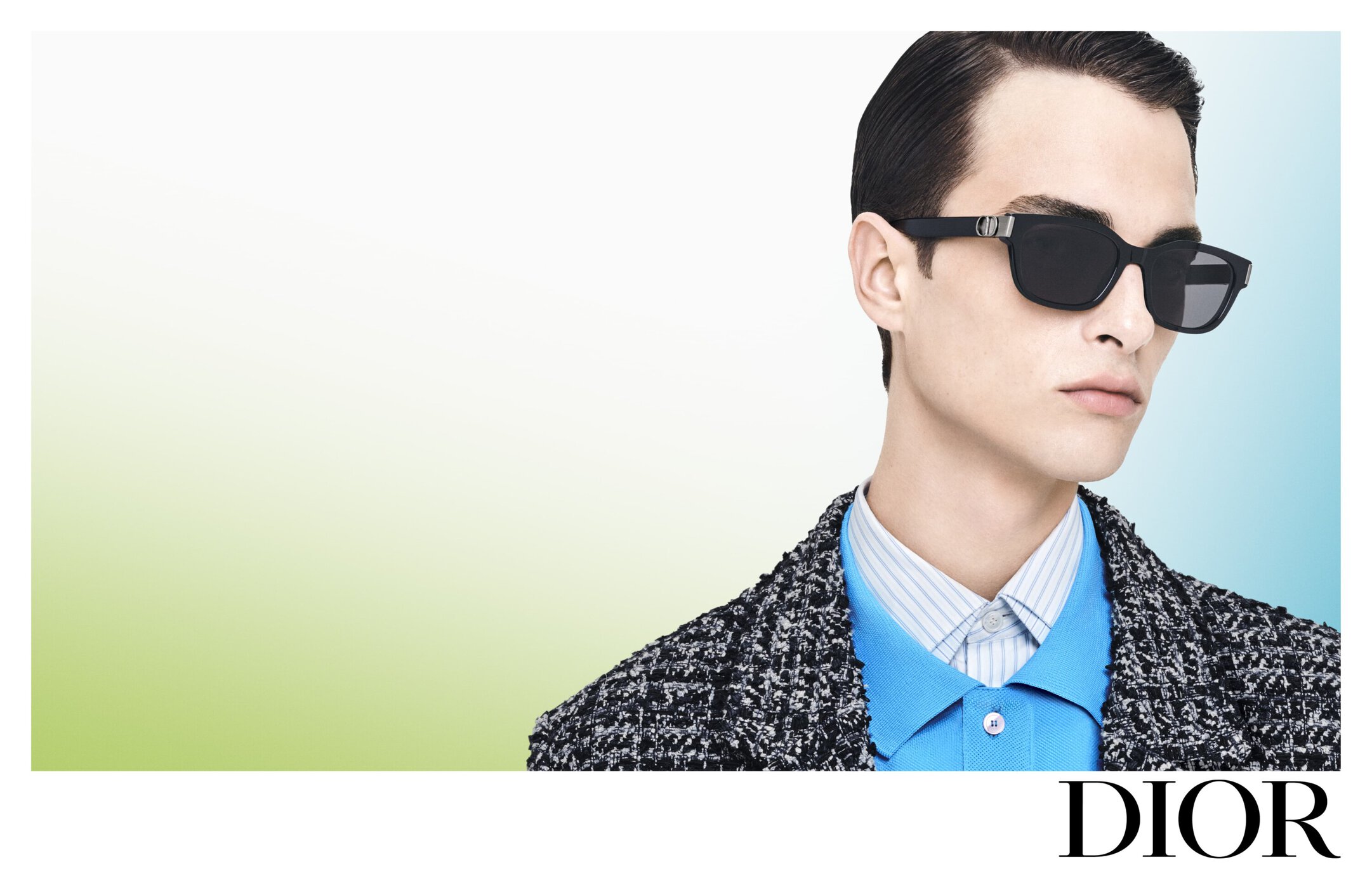 Fashion-forward portrait of a young adult man wearing stylish sunglasses, a patterned jacket, and a blue shirt against a gradient backdrop.