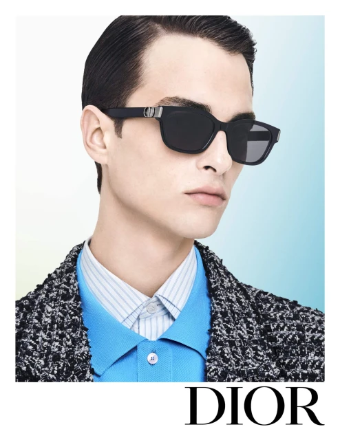 A close-up studio shot of a man modeling eyewear; wearing a patterned jacket, blue shirt, with a light gradient background.