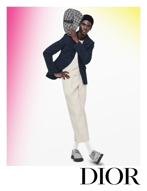An African American man is modeling a navy blazer, cream trousers, and patterned loafers, holding a handbag on his shoulder in a studio shot.