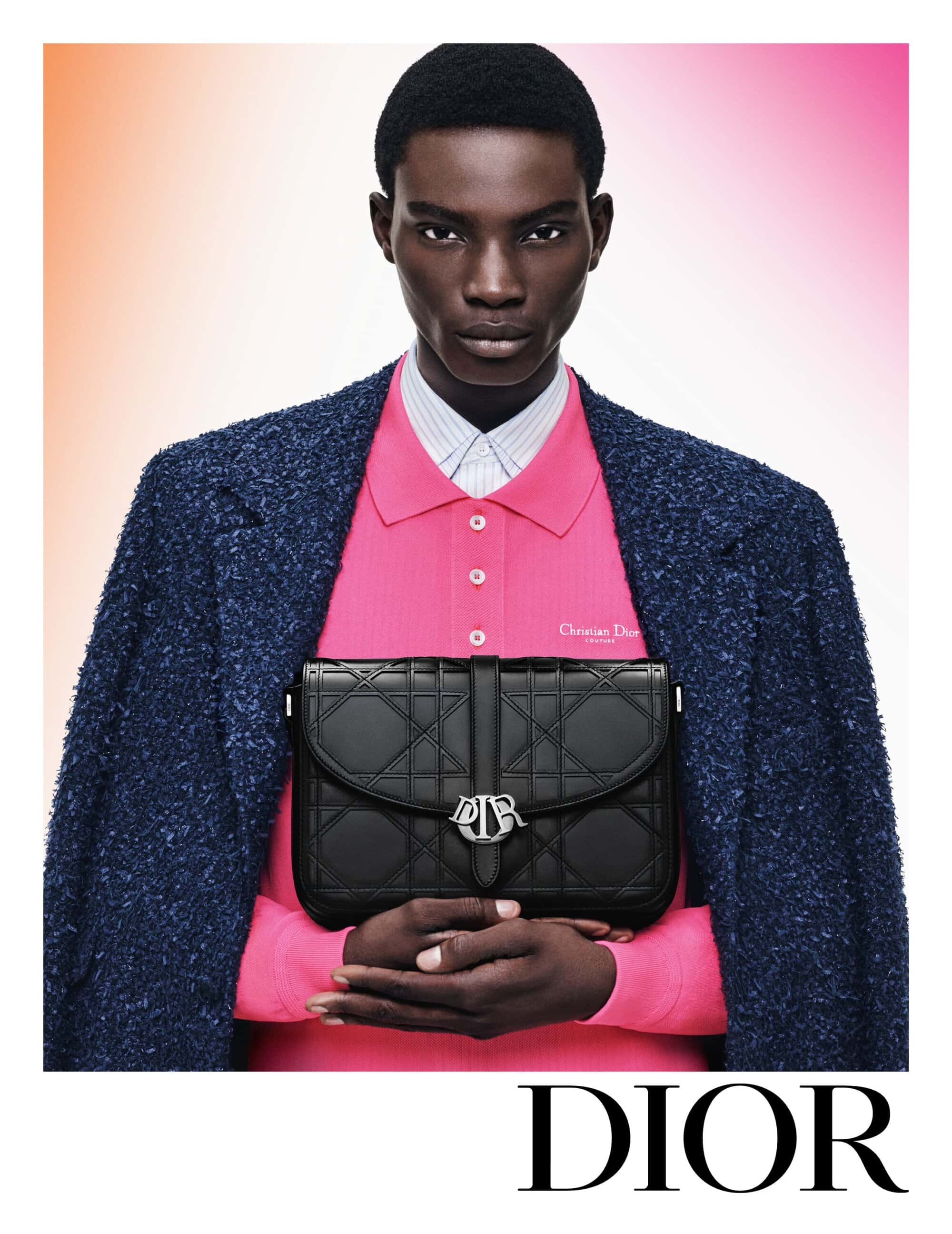 Fashion shot featuring a Black male model in a blue jacket and pink polo shirt, holding a black Dior handbag against a gradient background.