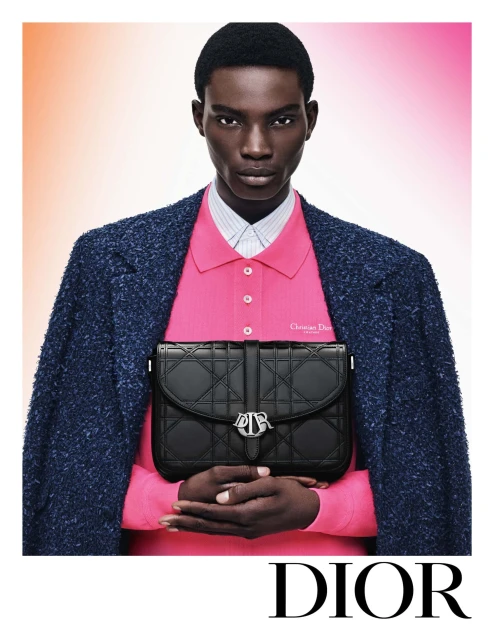 Fashion shot featuring a Black male model in a blue jacket and pink polo shirt, holding a black Dior handbag against a gradient background.