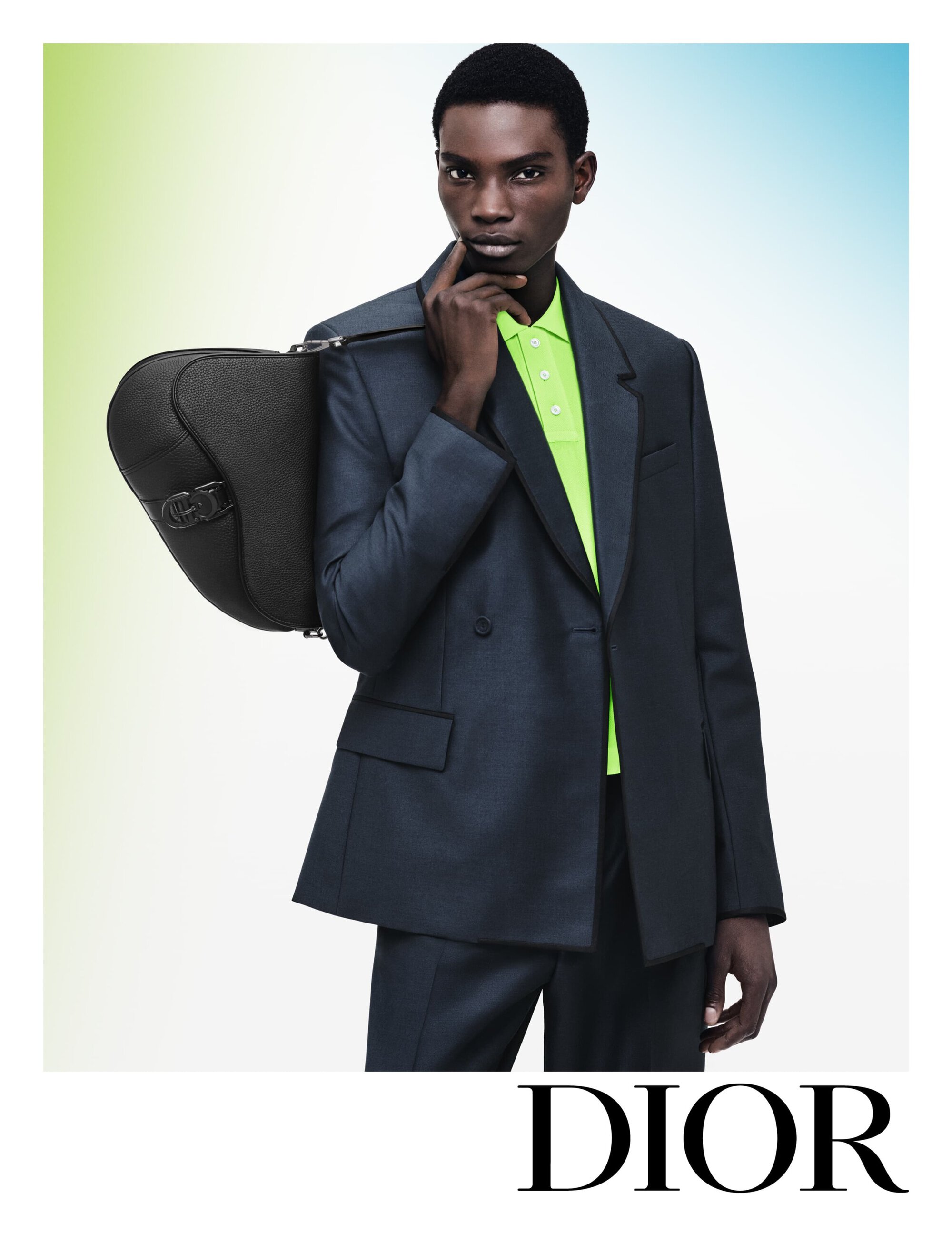 Elegant studio portrait of a Black male model wearing a dark suit and bright polo shirt, holding a designer handbag with logo.