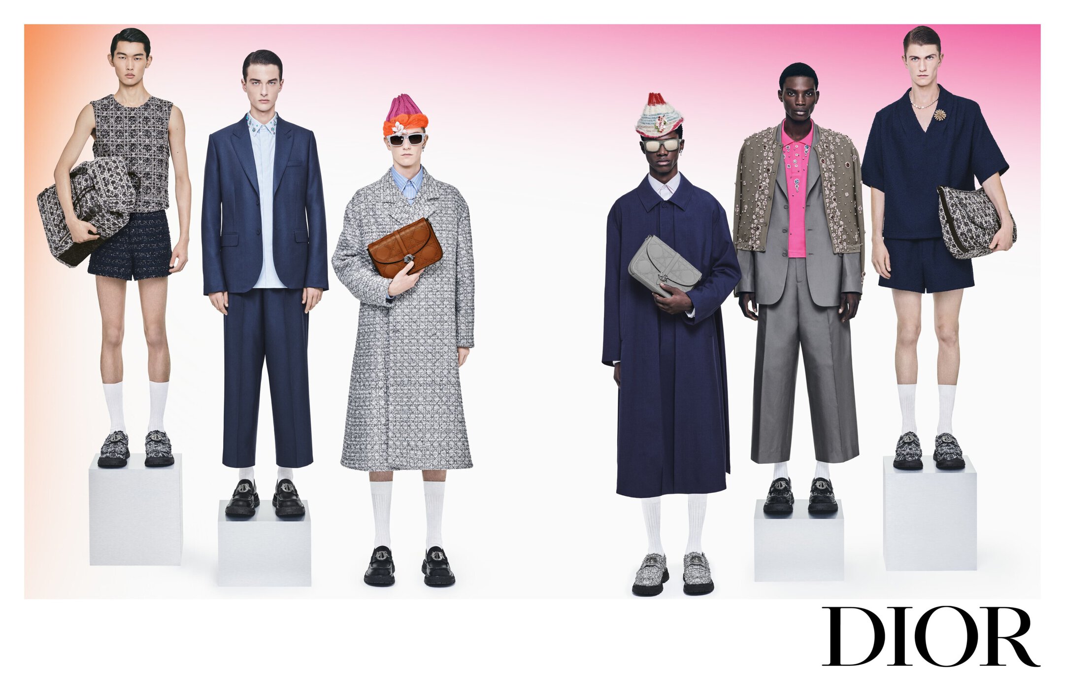 Fashion editorial shot of six male models in suits and separates, carrying patterned handbags, against a blended pink-and-orange backdrop.