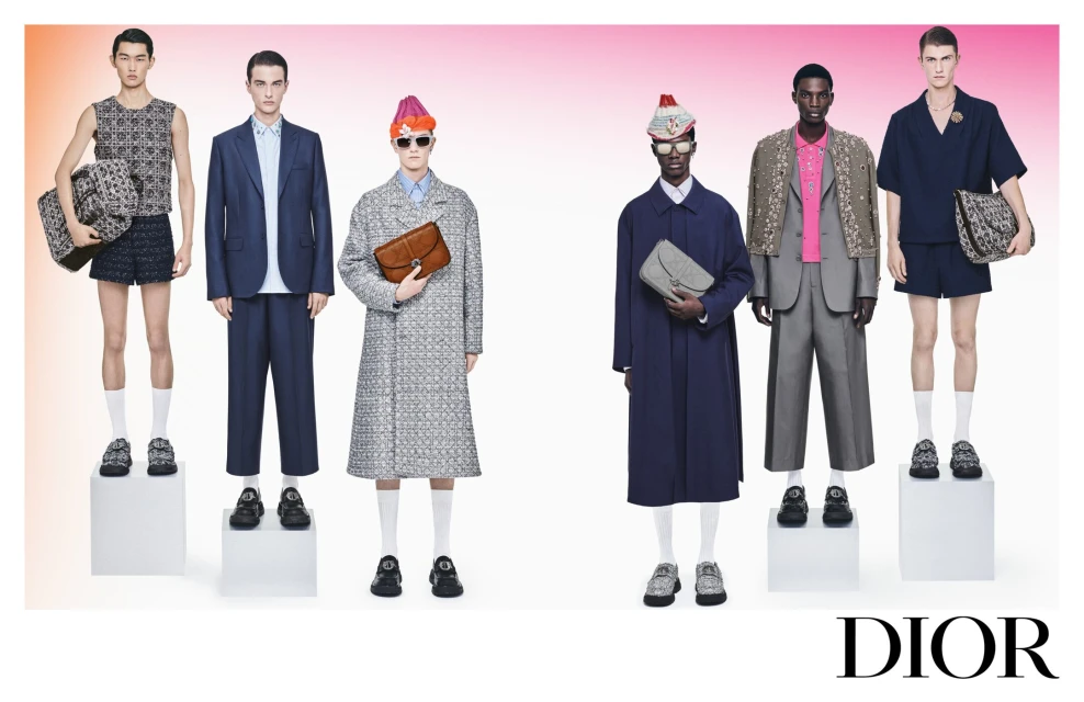 Fashion editorial shot of six male models in suits and separates, carrying patterned handbags, against a blended pink-and-orange backdrop.