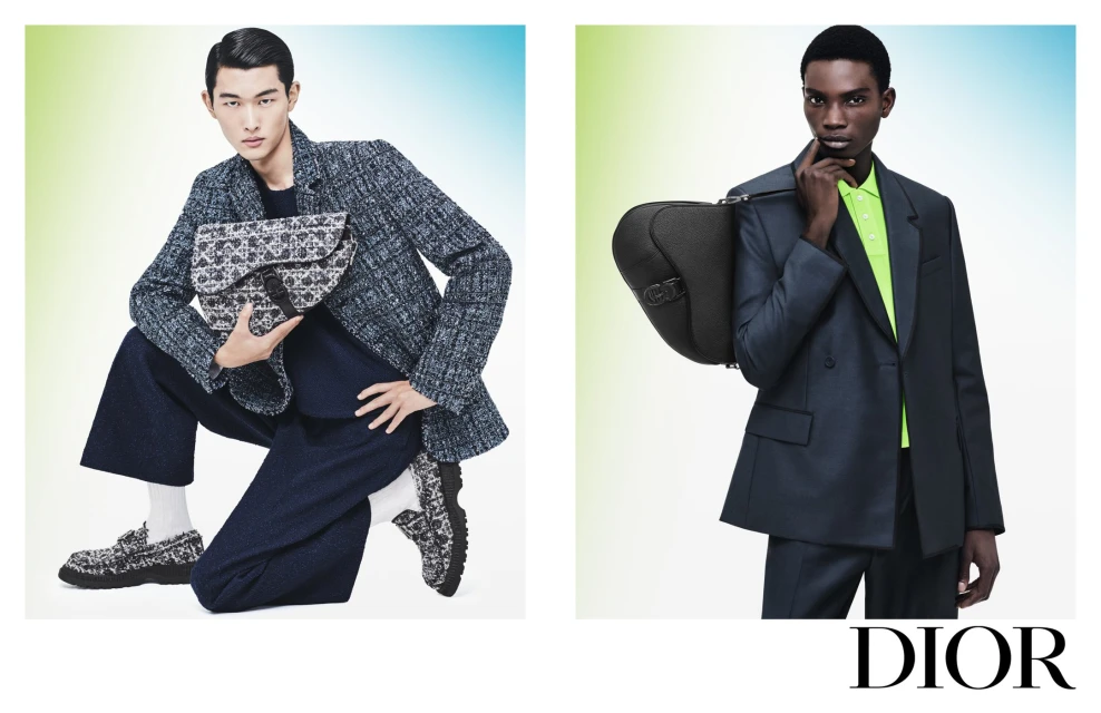 Two male fashion models in suits hold designer bags in a studio shot with a light gradient background, showcasing modern menswear.