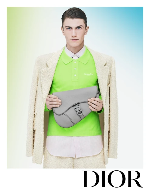 An adult male model poses in a photo studio wearing a light beige bouclé suit and a neon green polo shirt, holding a gray leather saddle bag.