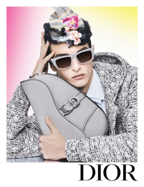 Fashion shot of a man wearing patterned sunglasses and a patterned jacket, holding a grey saddle bag, with pink and yellow background.