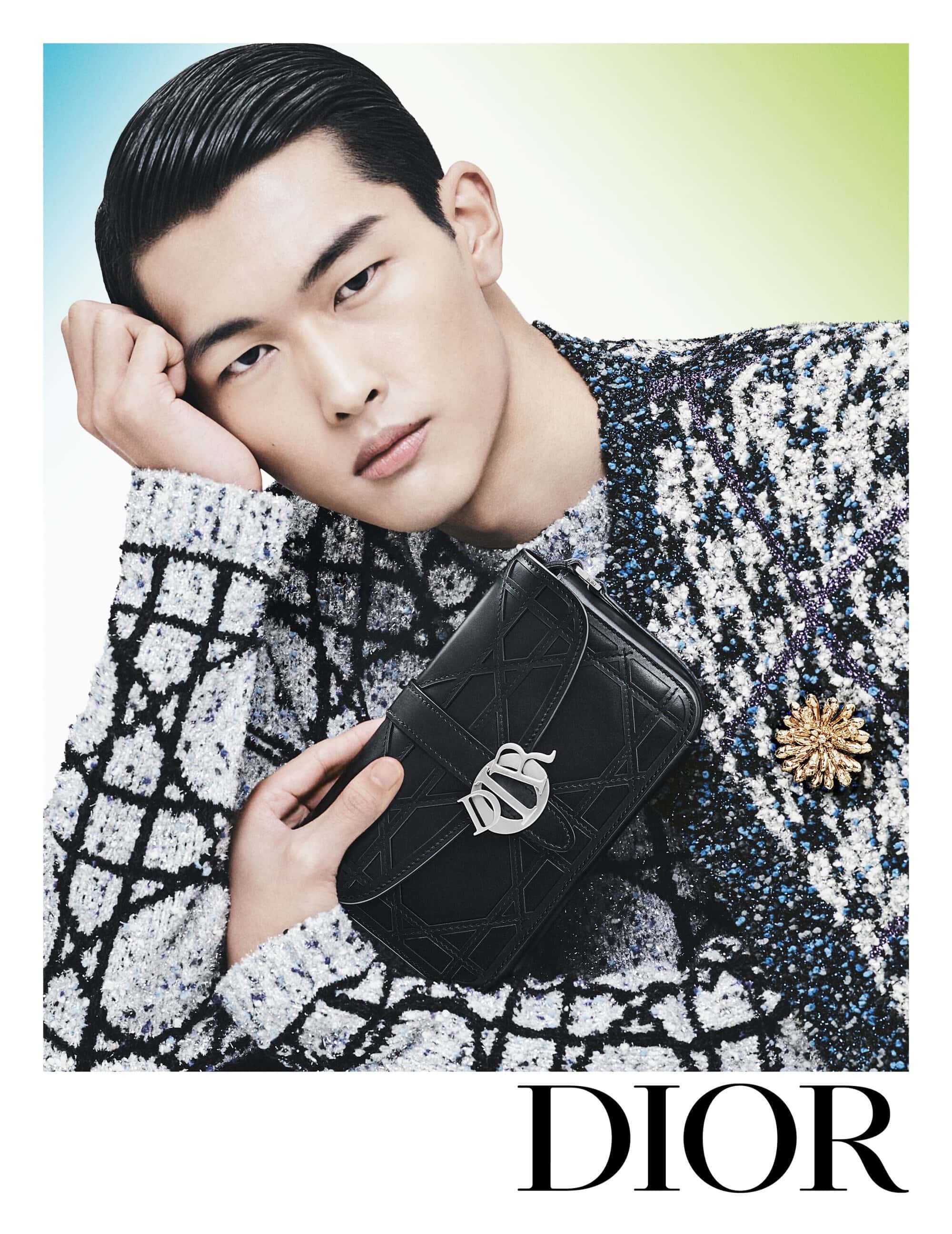 Asian male model poses with a black Dior purse and wearing a patterned sweater in a fashion studio with soft lighting.