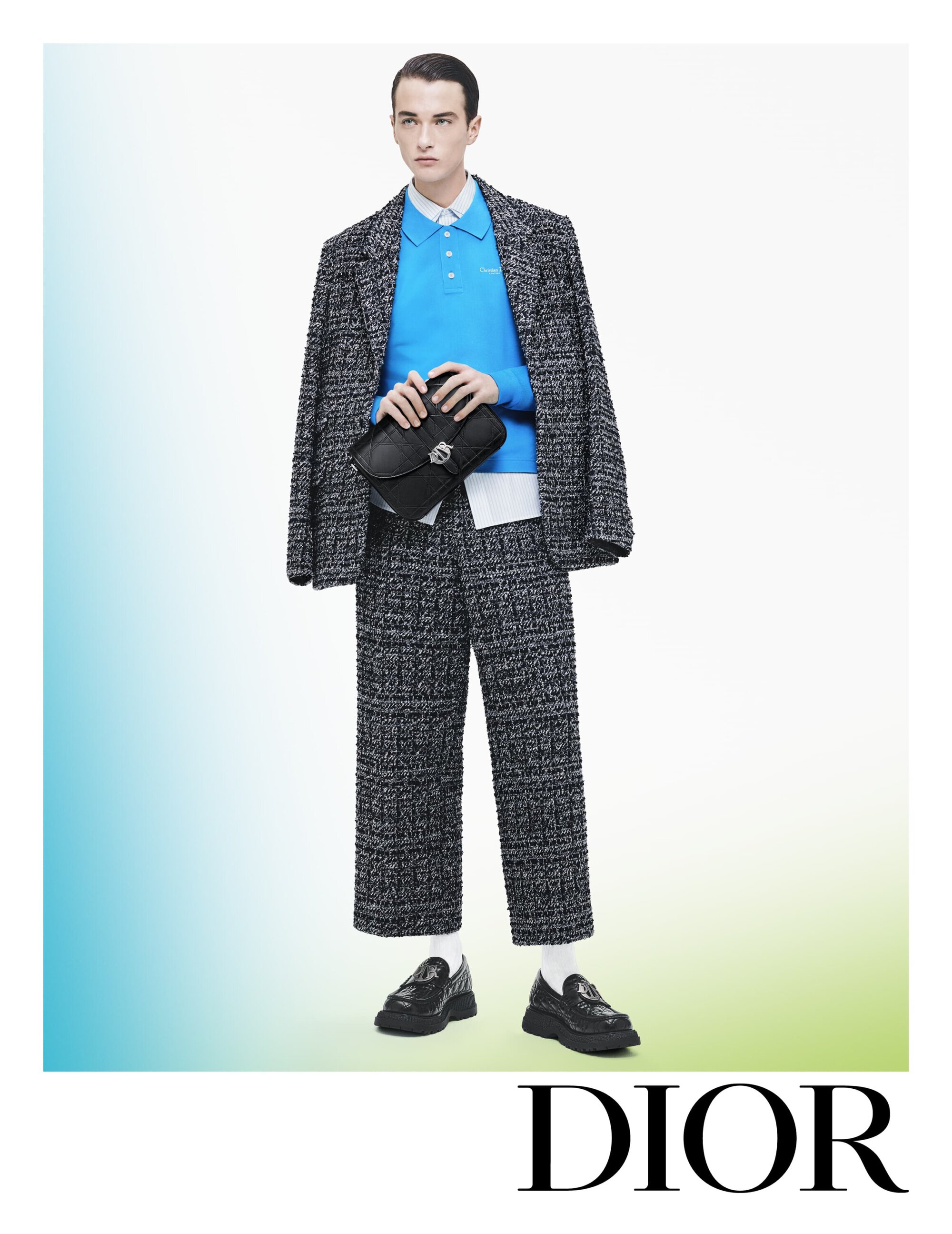 An adult male model poses in a bright studio wearing a grey speckled suit with a blue polo and black bag, with the brand name DIOR.